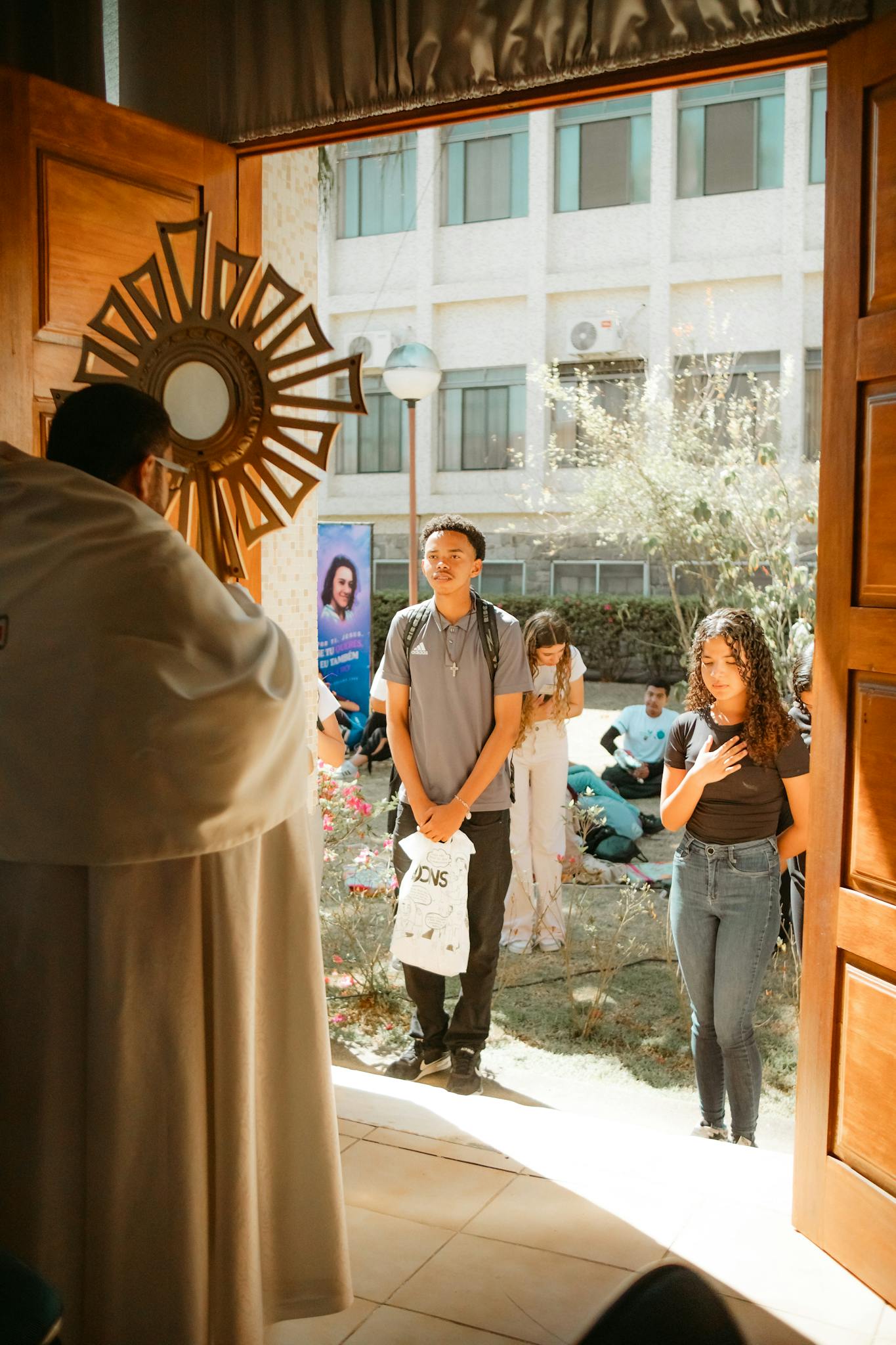A priest leads a religious ceremony with parishioners in a sunlit church entrance.